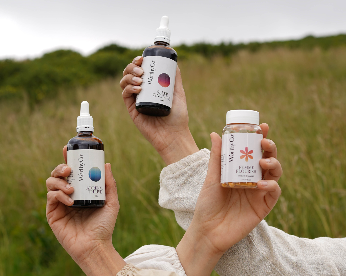 Three hands holding bottles of Worthy Co. products against a natural background
